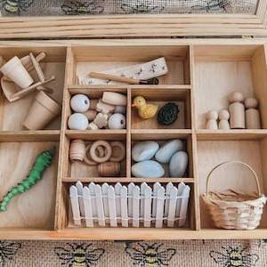Pu&ograve; includere: Set di giocattoli in legno con vari piccoli oggetti, tra cui un bruco verde, un'anatra gialla, perline di legno, una piccola staccionata bianca e un cestino in miniatura. Il set &egrave; disposto in una scatola di legno chiaro con pi&ugrave; scomparti.