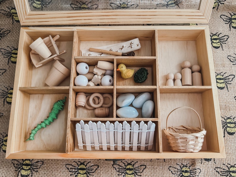 Pu&ograve; includere: Set di giocattoli in legno con vari piccoli oggetti, tra cui un bruco verde, un'anatra gialla, perline di legno, una piccola staccionata bianca e un cestino in miniatura. Il set &egrave; disposto in una scatola di legno chiaro con pi&ugrave; scomparti.