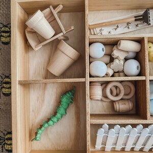 Pu&ograve; includere: Una scatola di legno con scomparti pieni di giocattoli di legno in miniatura. Include una carriola, vasi di fiori, perline, anelli, un bruco verde e una piccola staccionata bianca. La scatola &egrave; in legno chiaro.