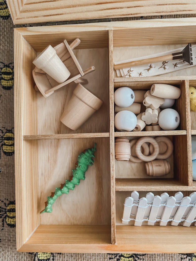 Pu&ograve; includere: Una scatola di legno con scomparti pieni di giocattoli di legno in miniatura. Include una carriola, vasi di fiori, perline, anelli, un bruco verde e una piccola staccionata bianca. La scatola &egrave; in legno chiaro.