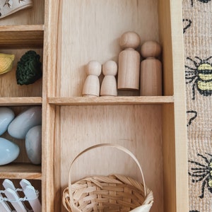 Pu&ograve; includere: Una scatola espositiva in legno con vari oggetti. Contiene bambole di legno, un cestino intrecciato e altri piccoli oggetti decorativi. La scatola &egrave; in legno chiaro e gli oggetti sono disposti in scomparti separati.