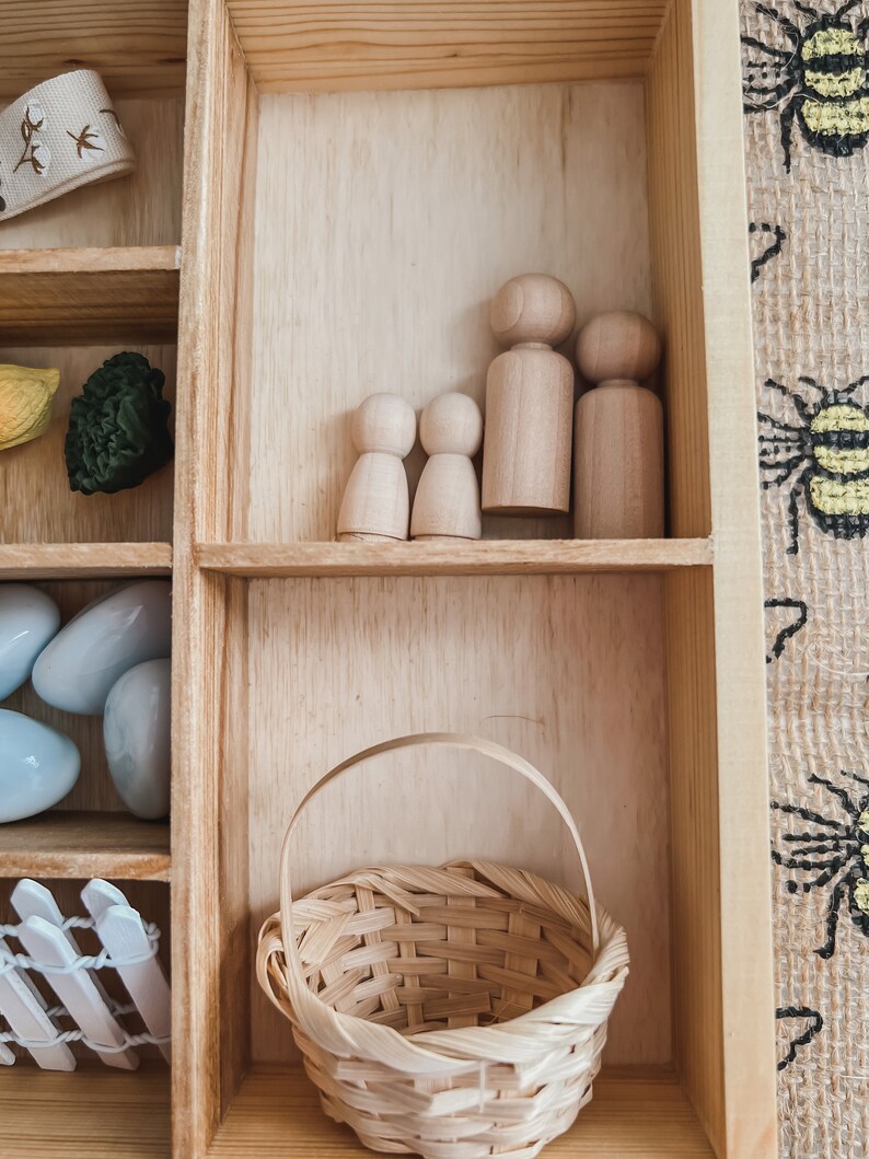 Pu&ograve; includere: Una scatola espositiva in legno con vari oggetti. Contiene bambole di legno, un cestino intrecciato e altri piccoli oggetti decorativi. La scatola &egrave; in legno chiaro e gli oggetti sono disposti in scomparti separati.