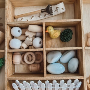 Pu&ograve; includere: Un organizer in legno con vari piccoli giocattoli in legno e plastica. Include un piccolo rastrello, perline bianche, barili di legno, anelli, un'anatra gialla, un cespuglio verde, pietre blu e una staccionata bianca.