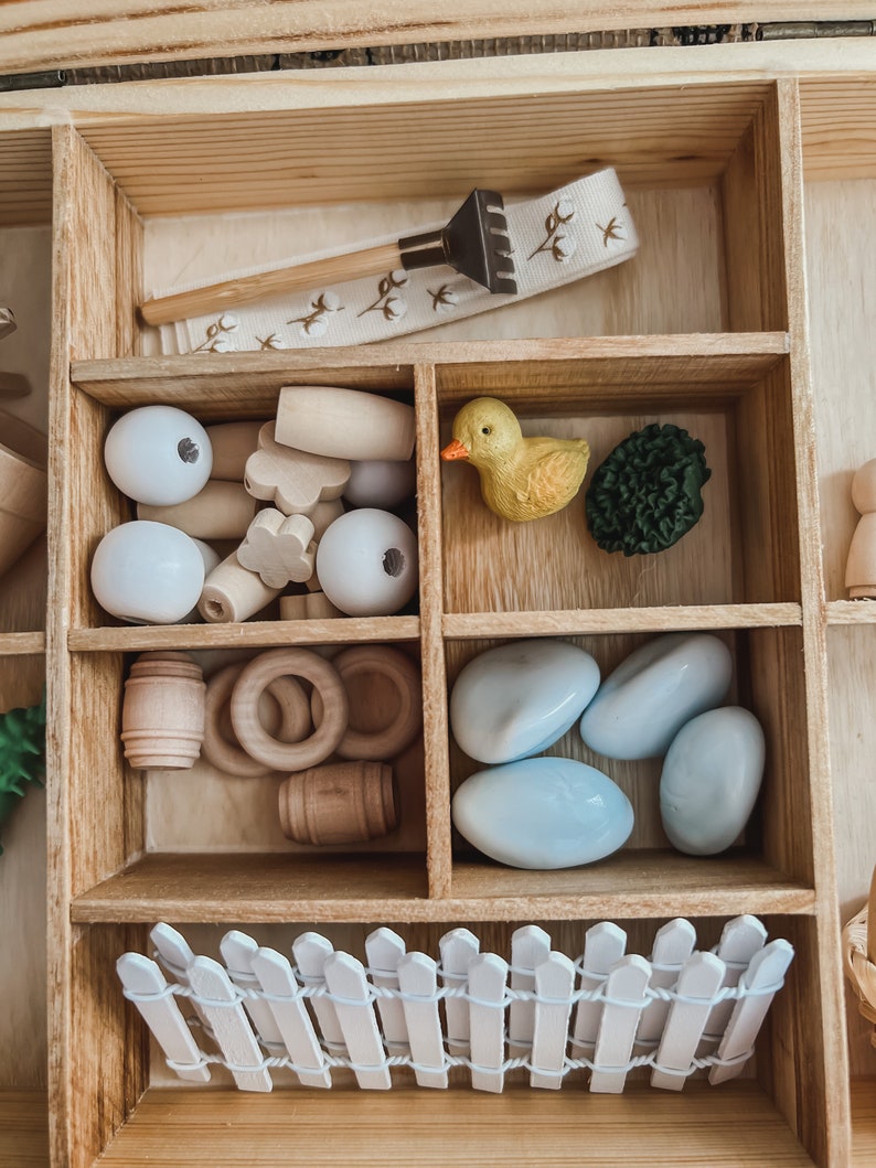 Pu&ograve; includere: Un organizer in legno con vari piccoli giocattoli in legno e plastica. Include un piccolo rastrello, perline bianche, barili di legno, anelli, un'anatra gialla, un cespuglio verde, pietre blu e una staccionata bianca.