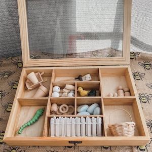 Pu&ograve; includere: Una scatola sensoriale in legno con coperchio trasparente, piena di giocattoli e accessori in legno in miniatura. Include una carriola, un'anatra, un bruco, una recinzione, un cestino e altri piccoli oggetti. La scatola &egrave; aperta, rivelando il contenuto.