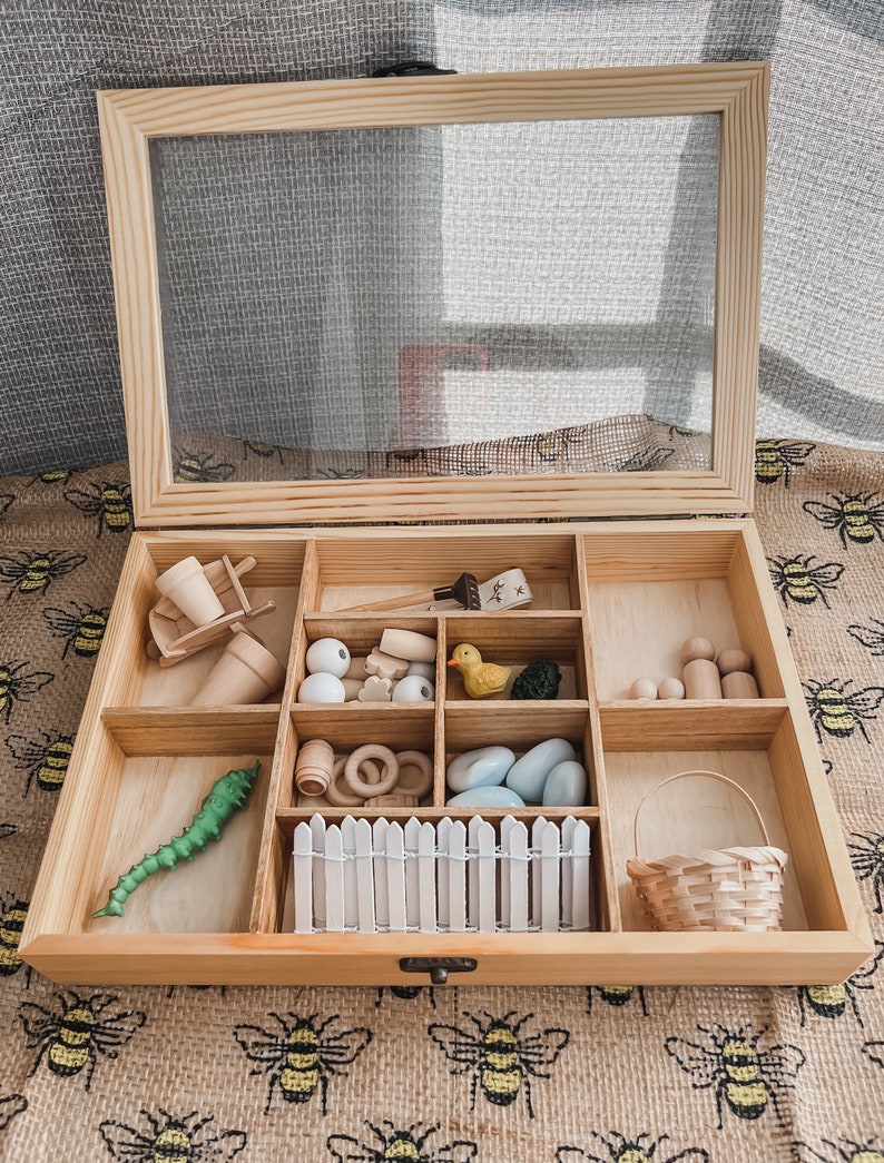 Pu&ograve; includere: Una scatola sensoriale in legno con coperchio trasparente, piena di giocattoli e accessori in legno in miniatura. Include una carriola, un'anatra, un bruco, una recinzione, un cestino e altri piccoli oggetti. La scatola &egrave; aperta, rivelando il contenuto.
