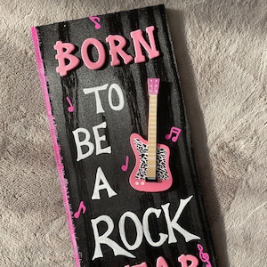 Peut inclure: Une pancarte en bois noir avec les mots "Born to be a Rock Star" en lettres roses et blanches. Une guitare rose avec un motif animal noir et blanc est fixée à la pancarte.