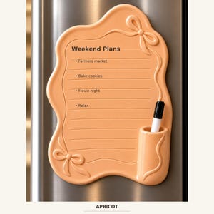 Puede incluir: Una pizarra magnética de borrado en seco color albaricoque con borde ondulado y dos lazos moldeados. La pizarra presenta el texto Weekend Plans, una lista con líneas y viñetas, y un soporte integrado con un marcador negro y blanco.