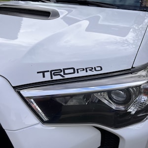 Puede incluir: Toyota 4Runner blanco con un emblema "TRD PRO" negro en el capó. El vehículo tiene un parachoques delantero negro con una moldura plateada y una parrilla negra.