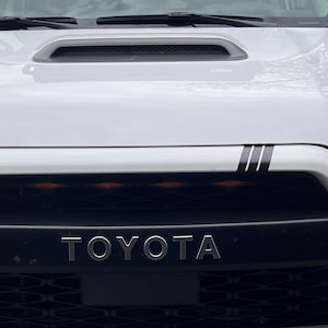 Könnte beinhalten: Ein weißer Toyota SUV mit einem schwarzen Kühlergrill und dem Schriftzug "TOYOTA" in silbernen Buchstaben. Das Fahrzeug hat schwarze Rennstreifen auf der Motorhaube.