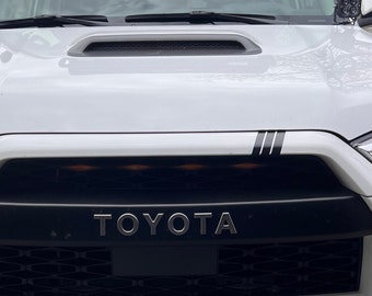 Può includere: Un SUV Toyota bianco con una griglia nera e la scritta "TOYOTA" in lettere argentate. Il veicolo ha delle strisce da corsa nere sul cofano.