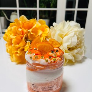 Peut inclure: Un pot en verre transparent rempli de slime orange et blanc en couches avec des tranches d'orange sur le dessus. Le pot porte une étiquette indiquant "Orange Cream Cloud Slime".
