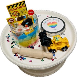 May include: A white ceramic cake stand with a yellow toy construction vehicle, a wooden rolling pin, and a glass jar filled with colorful play dough and various small toys. The jar has a label that reads "Sweetest Fidgets" with a rainbow heart.