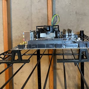 May include: A black computer motherboard with a silver heatsink and fan, resting on two black metal supports. The motherboard is on a black metal shelf.