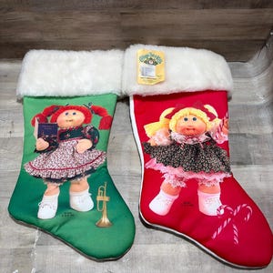 Könnte beinhalten: Zwei Weihnachtsstrümpfe mit weißem Kunstfellbesatz. Der grüne Strumpf zeigt eine Cabbage Patch Kids Puppe mit roten Zöpfen, die ein Buch und eine Trompete hält. Der rote Strumpf zeigt eine Puppe mit blondem Haar und ein Zuckerstangen-Motiv.