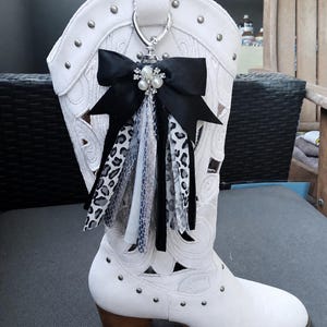 Può includere: Stivale da cowboy bianco con borchie decorative argentate e motivi traforati. Un fiocco nero con dettagli di perle e nastri fantasia pende da un anello argentato. Lo stivale ha un tacco in legno.