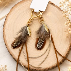 May include: A pair of gold-toned metal keychains featuring brown and grey patterned feathers, thin gold-toned curved metal accents, and brown suede-like leather cords, all attached to a gold-toned lobster clasp with a white rectangular display tag.