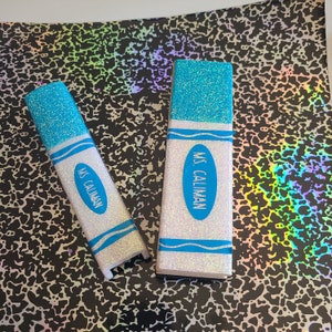 May include: Two white crayon-shaped pencil holders with blue glitter on the top and bottom. The holders have the text "MS. CALIMAN" printed on them.