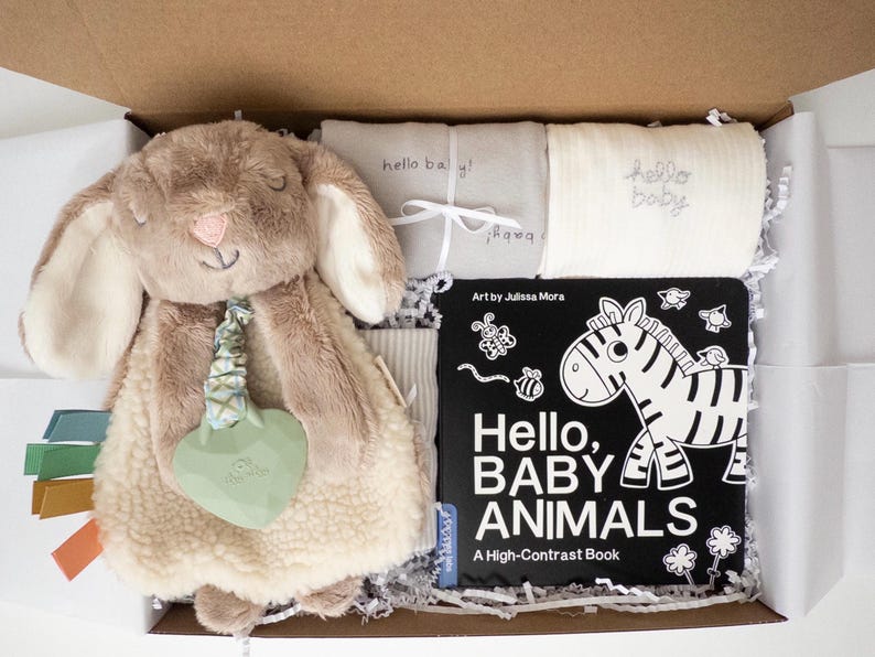 May include: A gift box containing a plush tan bunny security blanket with a green heart-shaped teether, a gray blanket with 'hello baby!' text, a white ribbed blanket with 'hello baby!' text, and a black and white 'Hello, BABY ANIMALS' book.