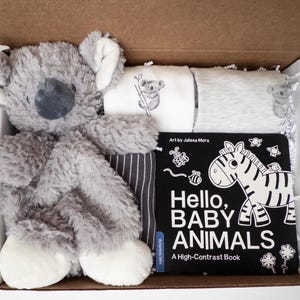 May include: A gift box containing a plush gray koala toy, two folded white and gray baby shirts with koala prints, and a black and white board book titled Hello, BABY ANIMALS A High-Contrast Book with a zebra illustration on the cover.