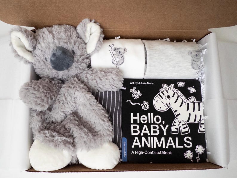May include: A gift box containing a plush gray koala toy, two folded white and gray baby shirts with koala prints, and a black and white board book titled Hello, BABY ANIMALS A High-Contrast Book with a zebra illustration on the cover.