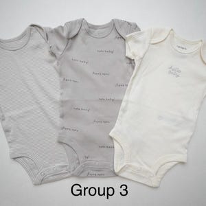 May include: Three short-sleeved baby bodysuits. The left one is light gray with thin horizontal stripes. The center one is medium gray with the repeating text hello baby!. The right one is cream with the text hello baby! embroidered.