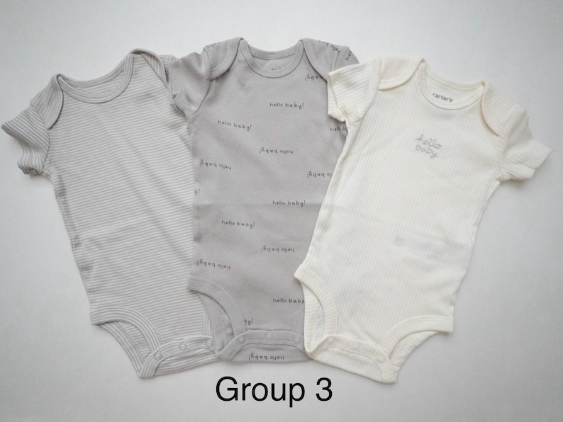 May include: Three short-sleeved baby bodysuits. The left one is light gray with thin horizontal stripes. The center one is medium gray with the repeating text hello baby!. The right one is cream with the text hello baby! embroidered.