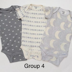 May include: Three short-sleeve baby bodysuits labeled Group 4. The left is dark gray with white rainbows. The center is cream with gray moon phases. The right is light gray with large cream crescent moons. Each has a Gerber brand tag.