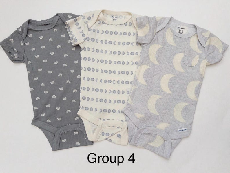 May include: Three short-sleeve baby bodysuits labeled Group 4. The left is dark gray with white rainbows. The center is cream with gray moon phases. The right is light gray with large cream crescent moons. Each has a Gerber brand tag.