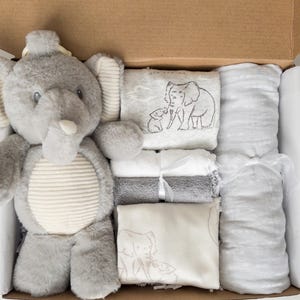 May include: A gift box containing a plush gray elephant toy with a ribbed belly, a folded white blanket with a line-art elephant print, two small rolled towels in white and gray, and a long, rolled light gray textured fabric wrap.