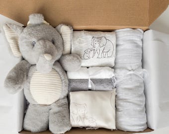 May include: A gift box containing a plush gray elephant toy with a cream-colored ribbed belly, two folded blankets featuring elephant illustrations, a white and a gray washcloth tied with ribbon, and a long, rolled gray patterned blanket.