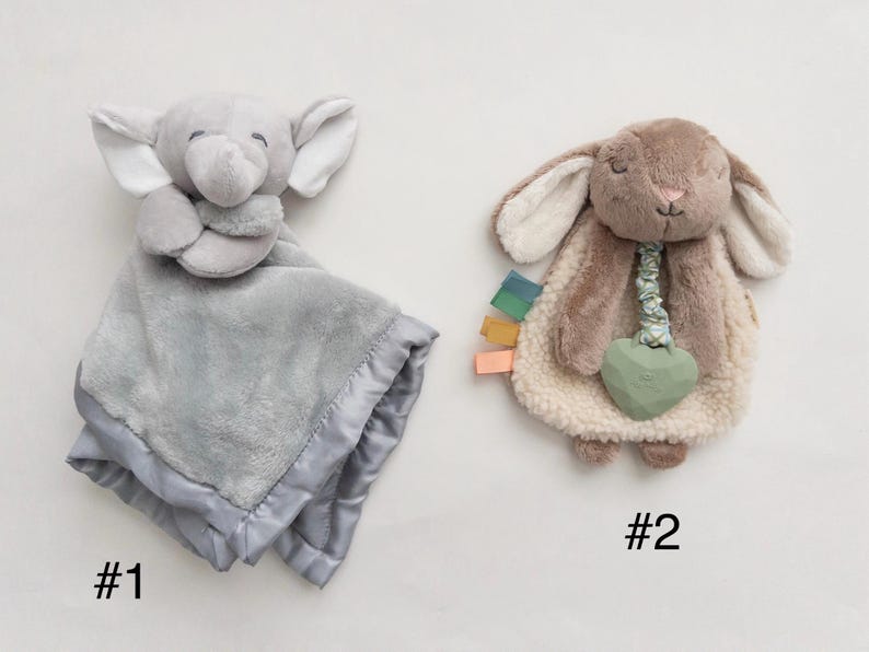 May include: Two plush security blankets. Number 1 is a gray elephant with a soft fabric body and satin trim. Number 2 is a brown rabbit with a textured cream-colored body, a green heart-shaped teether, and small colorful fabric tags on the side.