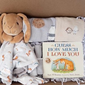 May include: A gift box containing a brown plush rabbit security blanket with a moon and star pattern, a gray shirt with a cookie and milk graphic, a striped shirt with a cookie and milk illustration, and a Guess How Much I Love You board book.