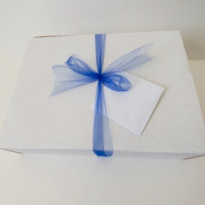 Puede incluir: Una caja rectangular de cart&oacute;n blanco envuelta con una cinta de tul azul vibrante atada en un lazo en el centro. Un sobre cuadrado blanco liso est&aacute; metido cuidadosamente debajo de la cinta de tul azul en la parte superior de la caja.