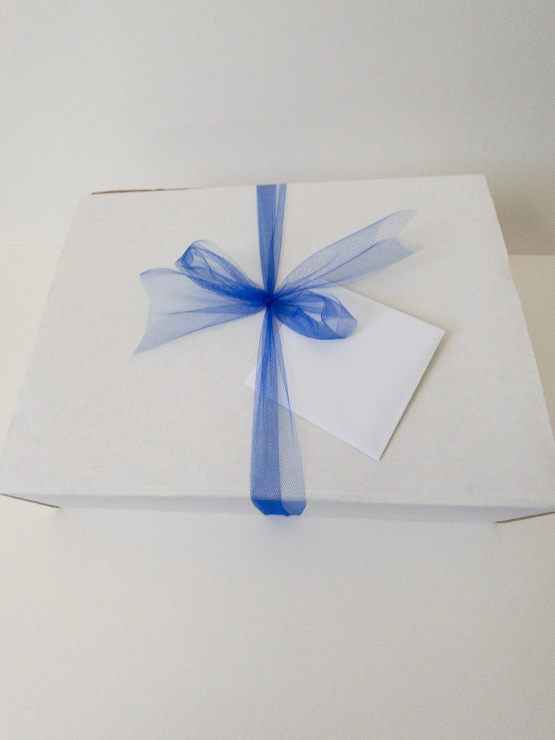 Puede incluir: Una caja rectangular de cart&oacute;n blanco envuelta con una cinta de tul azul vibrante atada en un lazo en el centro. Un sobre cuadrado blanco liso est&aacute; metido cuidadosamente debajo de la cinta de tul azul en la parte superior de la caja.