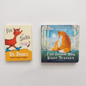 Puede incluir: Dos libros de cart&oacute;n con portadas ilustradas. El libro de la izquierda se titula Fox in Socks de Dr. Seuss, mostrando un zorro rojo con calcetines azules. El libro de la derecha se titula I've Loved You Since Forever de Hoda Kotb, representando a dos zorros en un bosque.