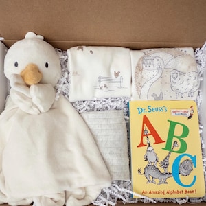 May include: A gift box containing a cream-colored plush duck security blanket, two folded white cloths with farm-themed illustrations, a gray textured fabric square, and a Dr. Seuss's ABC An Amazing Alphabet Book board book.