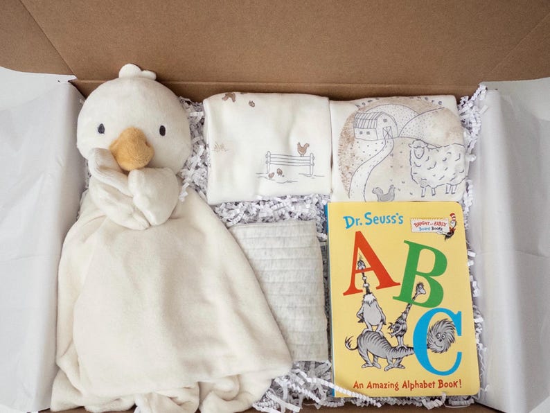May include: A gift box containing a cream-colored plush duck security blanket, two folded white cloths with farm-themed illustrations, a gray textured fabric square, and a Dr. Seuss's ABC An Amazing Alphabet Book board book.