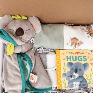 May include: A baby gift set in a box, featuring a gray koala plush toy with a teal blanket, a book titled "Babies Love HUGS", and other baby items. The book has a koala illustration.