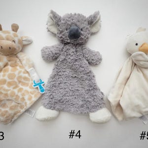 May include: Three plush security blankets featuring animal heads: a tan and white giraffe with a blue pacifier clip, a gray koala with textured fur, and a cream-colored duck. Each soft toy has a flat, blanket-like body for comfort.