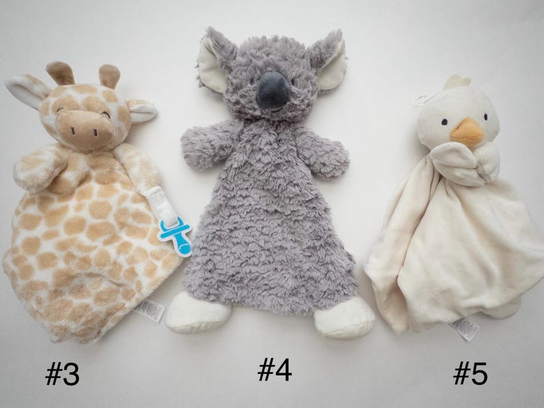 May include: Three plush security blankets featuring animal heads: a tan and white giraffe with a blue pacifier clip, a gray koala with textured fur, and a cream-colored duck. Each soft toy has a flat, blanket-like body for comfort.