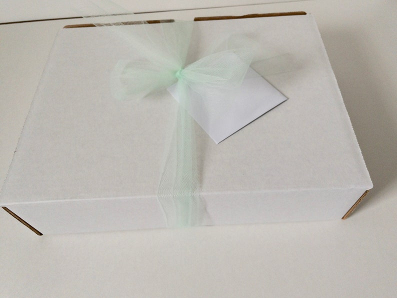 May include: A rectangular white cardboard box tied with a pale mint green tulle ribbon. A small, blank, square white card is tucked under the ribbon bow on the top surface of the box. The box is closed and features a clean, simple design.