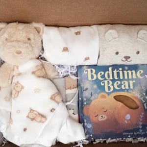 May include: A gift box containing a plush tan teddy bear attached to a white security blanket with bear patterns, a white onesie with small bear prints, a beige bear-faced hat, and a Bedtime Bear board book with a brown bear illustration.