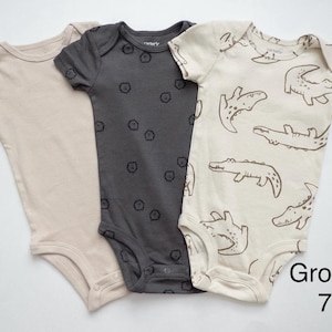 May include: Three short-sleeve baby bodysuits with snap closures at the bottom. One is solid beige, one is dark gray with small black lion icons, and one is cream with a brown alligator print. The text Group 7 is visible in the corner.