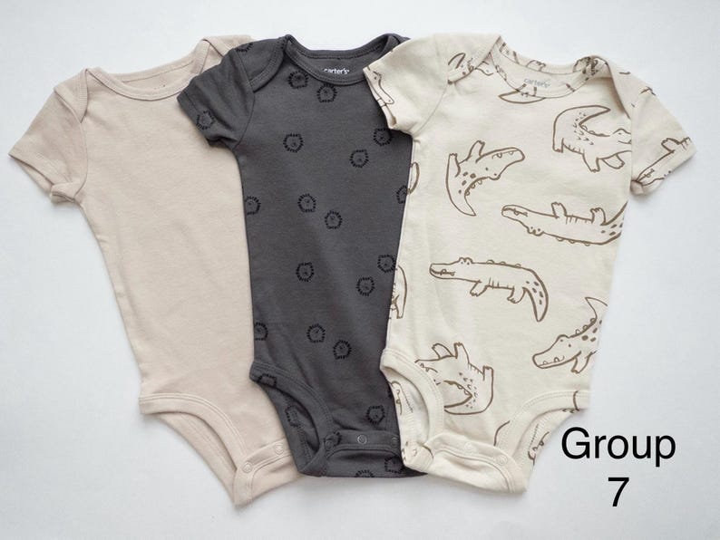 May include: Three short-sleeve baby bodysuits with snap closures at the bottom. One is solid beige, one is dark gray with small black lion icons, and one is cream with a brown alligator print. The text Group 7 is visible in the corner.