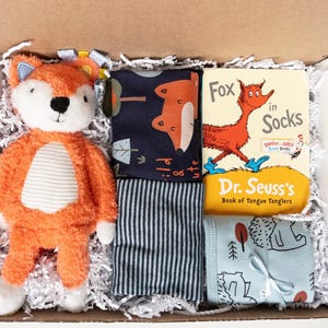 Puede incluir: Una caja de regalo que contiene un peluche de zorro naranja y blanco, una tela azul oscuro con estampado de zorros, una tela a rayas grises y blancas, una tela azul claro con estampado de erizos y el libro Fox in Socks de Dr. Seuss.