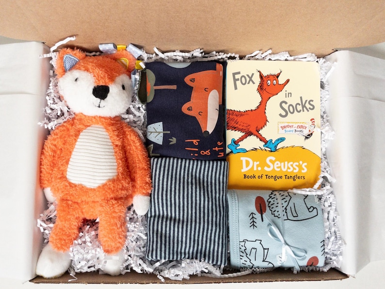 Puede incluir: Una caja de regalo que contiene un peluche de zorro naranja y blanco, una tela azul oscuro con estampado de zorros, una tela a rayas grises y blancas, una tela azul claro con estampado de erizos y el libro Fox in Socks de Dr. Seuss.