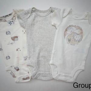 May include: Three short-sleeved baby bodysuits. The left one is cream with a brown farm animal print. The center one is a solid light gray ribbed knit. The right one is white with a circular graphic of a farm and the text Home is where my mama is.