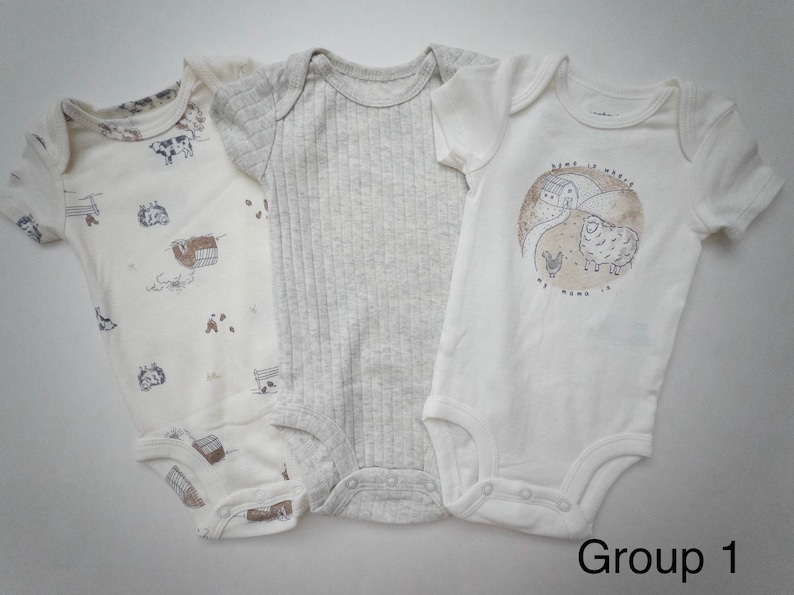 May include: Three short-sleeved baby bodysuits. The left one is cream with a brown farm animal print. The center one is a solid light gray ribbed knit. The right one is white with a circular graphic of a farm and the text Home is where my mama is.