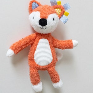 Puede incluir: Un peluche de zorro con pelaje naranja texturizado, cara blanca y vientre blanco con rayas horizontales. El zorro tiene una nariz negra, patas blancas y cintas azules y amarillas unidas a una oreja. Las orejas tienen detalles en azul y amarillo.
